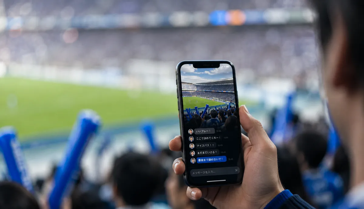 スタジアムの観客席でスマートフォンを掲げ、試合の映像の下にファンの応援コメントが次々と流れる応援アプリの画面を見ている様子